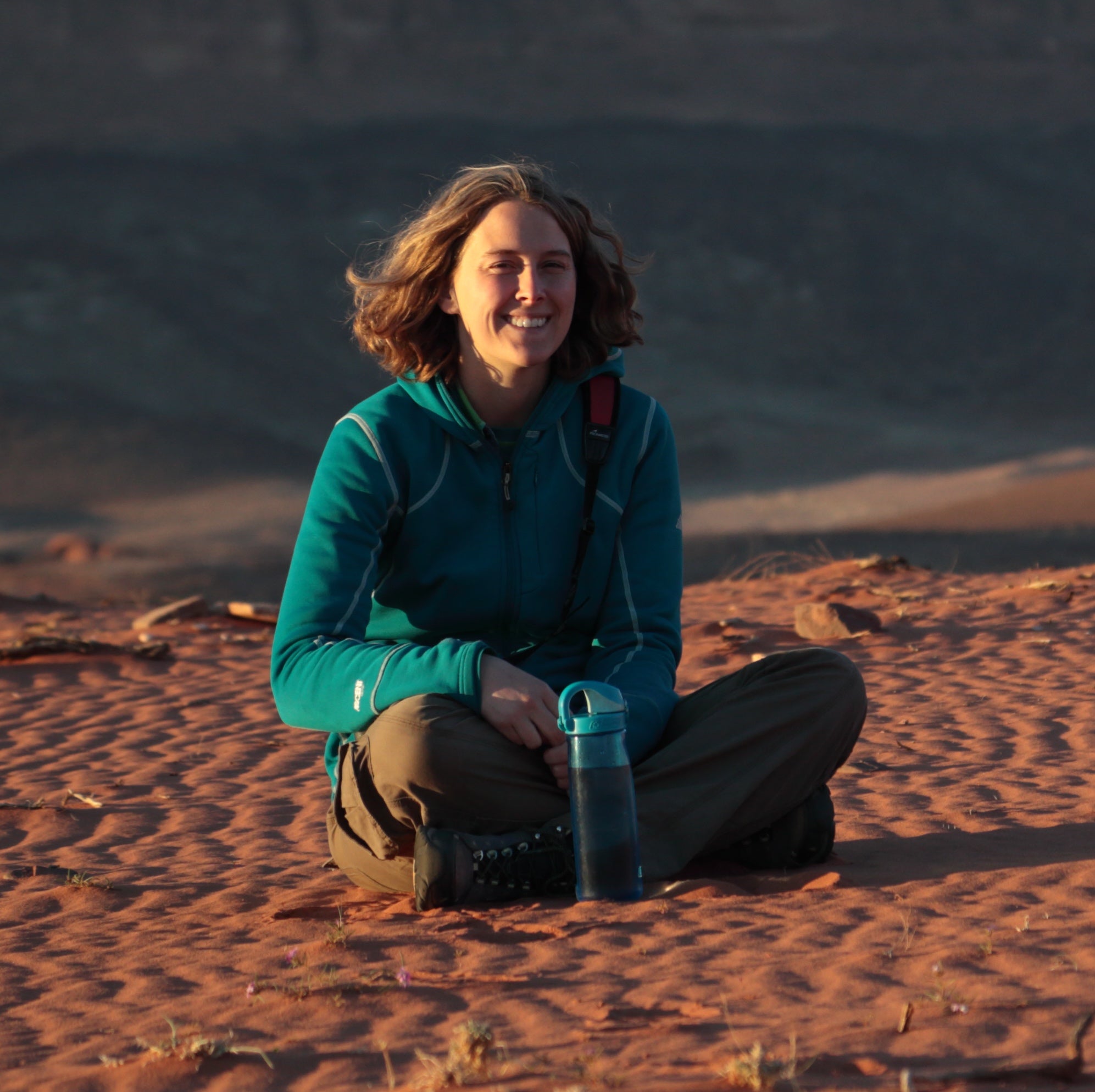 Jessica sits cross-legged in a field of desert sand, a water bottle in front of her and a smile on her face. 