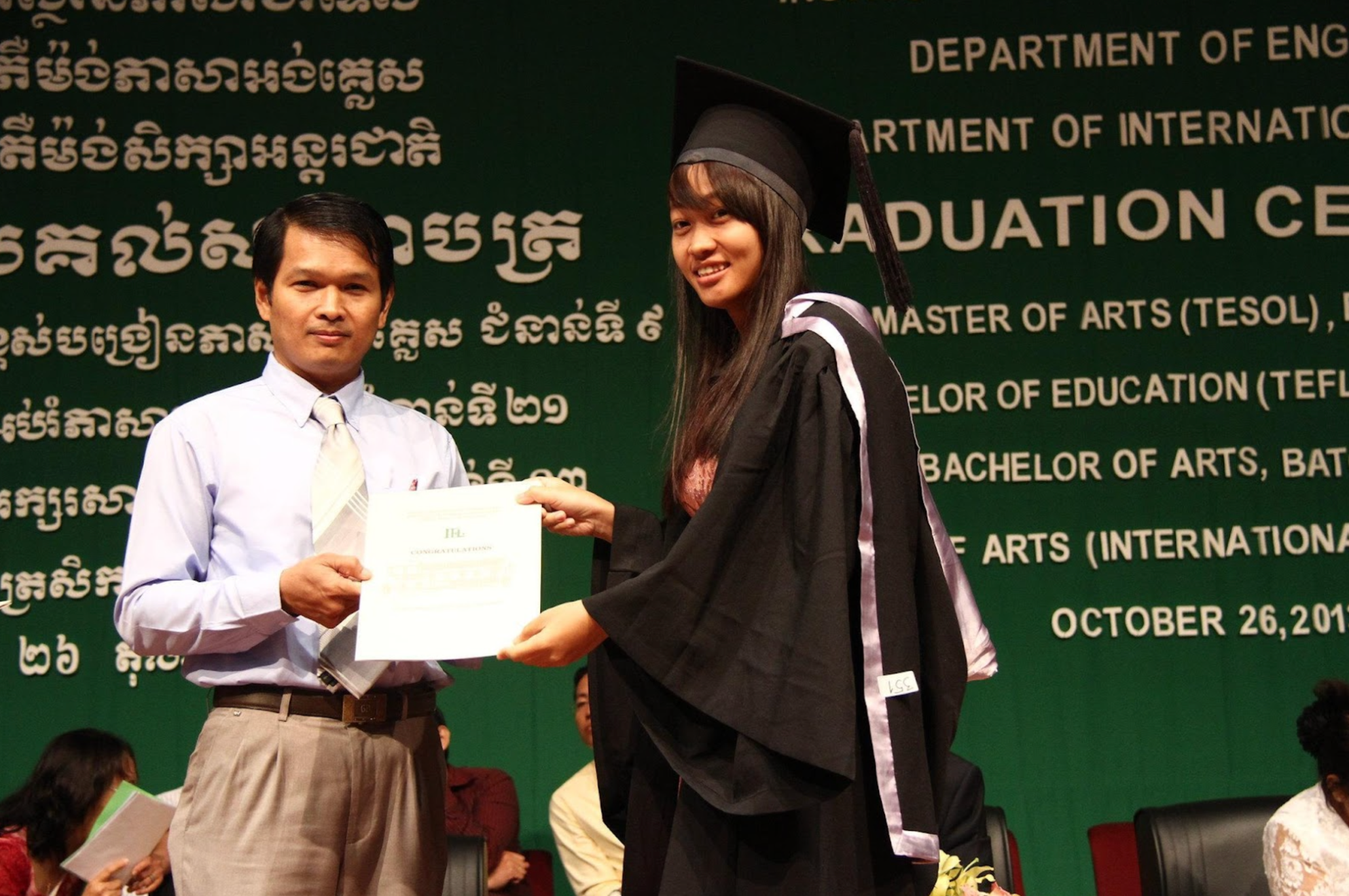 An official and a graduate are holding a certificate together against a green background that reads “Graduation Ceremony” and “Master of Arts (TESOL)”