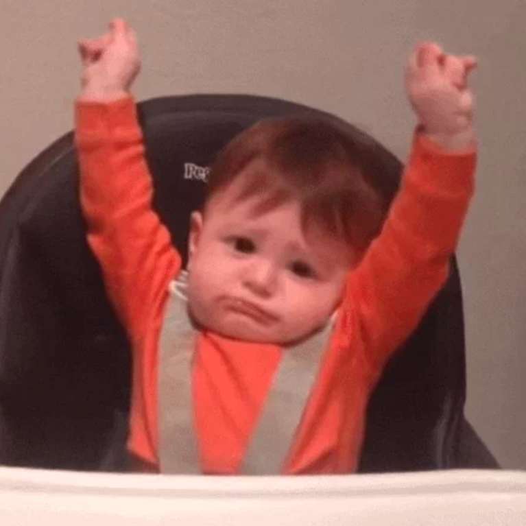 A seated baby strapped into his high chair stretches both arms up. 