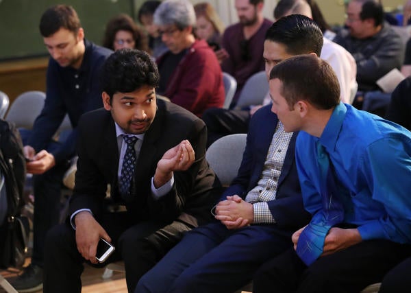 Three young men have a spirited conversation while seated in a row at an event. 