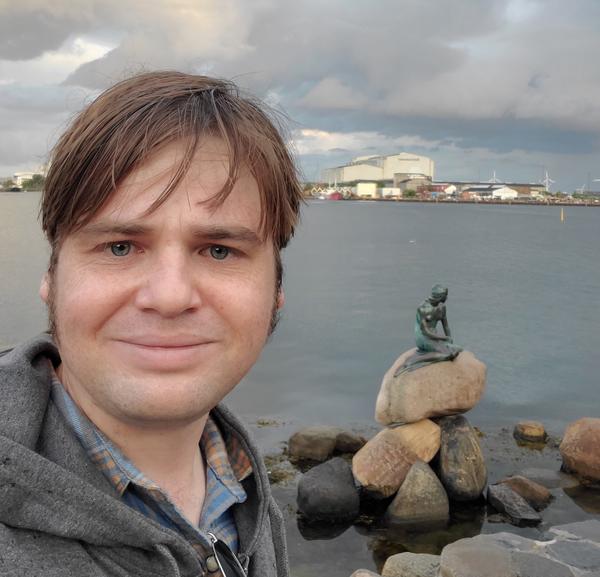 Bryan from the shoulders up, standing in front of the Hans Christian Anderson mermaid statue in Copenhagen harbor.