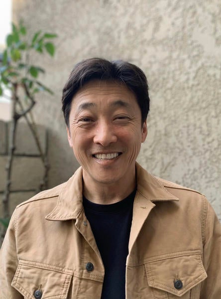 A photo of Jason from the shoulders up, smiling, wearing a black t-shirt and tan jacket