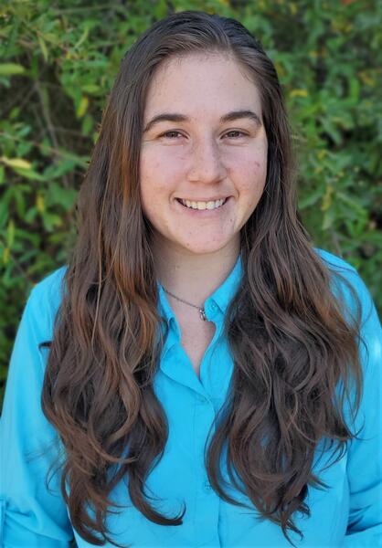 Samantha from mid-torso up, wearing a bright blue shirt and smiling in front of a green bush