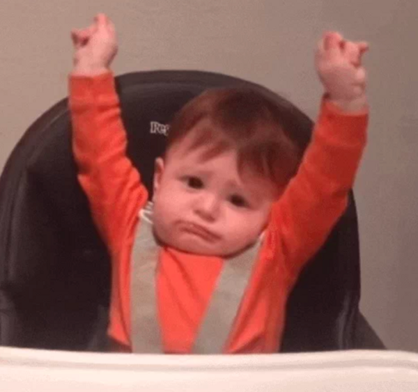 A seated baby strapped into his high chair stretches both arms up. 