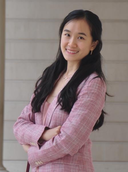 Photo of Yi from waist up in a pink jacket with arms crossed, smiling