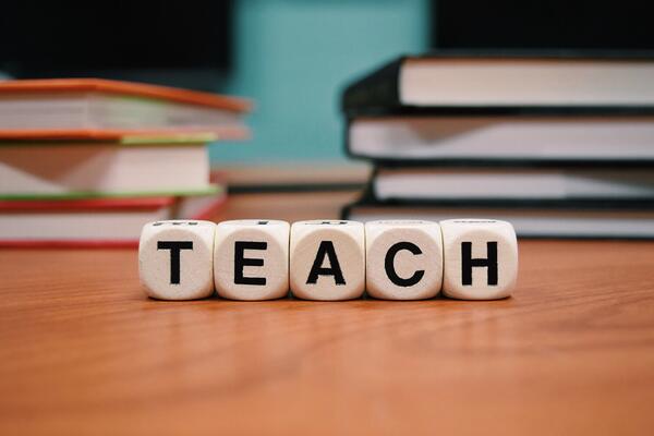 Four dice are aligned to spell the word "TEACH" on a desk with blurred books in the background.